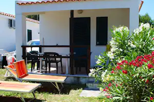 Terrasse am Campinghaus mit zwei Liegestühlen, Holztisch und Stühlen, umgeben von blühenden Sträuchern und weißer Fassade.