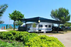 Wohnmobil auf dem Stellplatz unter einer Segelmarkise, Hecken und Bäume am Weg, Meer im Hintergrund.
