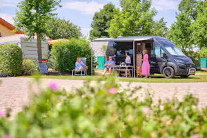 Wohnmobil auf dem Campingplatz mit Sitzgruppe und mehreren Personen vor dem Fahrzeug, umgeben von Bäumen und Hecken.