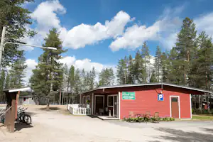 Rotes Campinggebäude mit Eingang, Terrasse und Sitzbänken im Wald, daneben Fahrräder und ein Parkplatzschild.