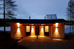 Holzhütte am See mit beleuchteter Holzterrasse, zwei Türen und Fensterfront, im Hintergrund Bäume und dunkler Himmel.
