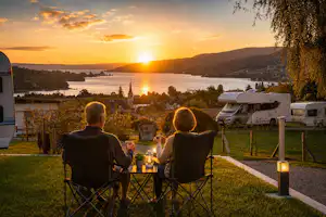 Zwei Personen sitzen abends an einem Tisch mit Kerzen auf dem Campingplatz, im Hintergrund Wohnmobile und eine Flusslandschaft.