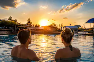 Zwei Personen im Pool mit Blick auf Campingplatz, Wohnwagen und Zelte bei Sonnenuntergang, umgeben von Bäumen und Stegen.