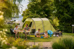 Campingzelt im Garten mit Tisch und Stühlen, Kinder spielen am Boden, im Hintergrund Bäume und ein Seeufer.