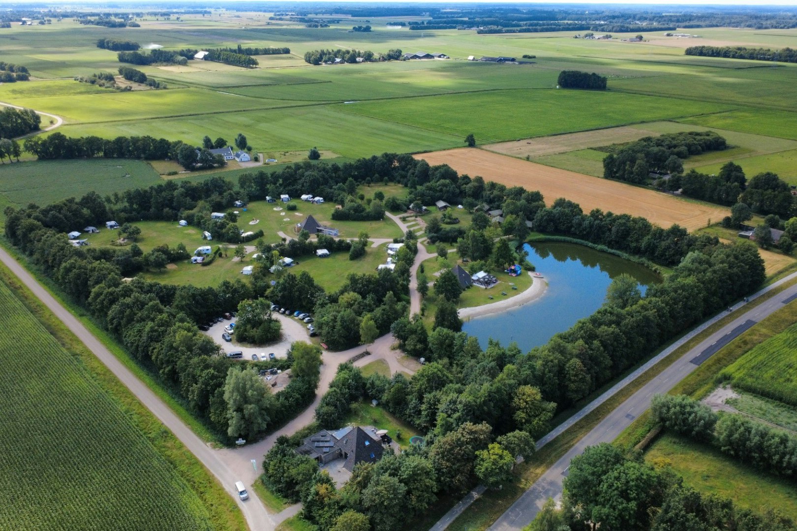Luftaufnahme eines Campingplatzes mit Wohnwagen und Zelten, umgeben von Bäumen, Wegen und einem kleinen See im Grünen.