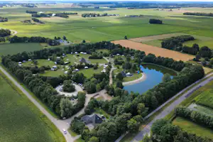 Luftaufnahme eines Campingplatzes mit Wohnwagen und Zelten, umgeben von Bäumen, Wegen und einem kleinen See im Grünen.