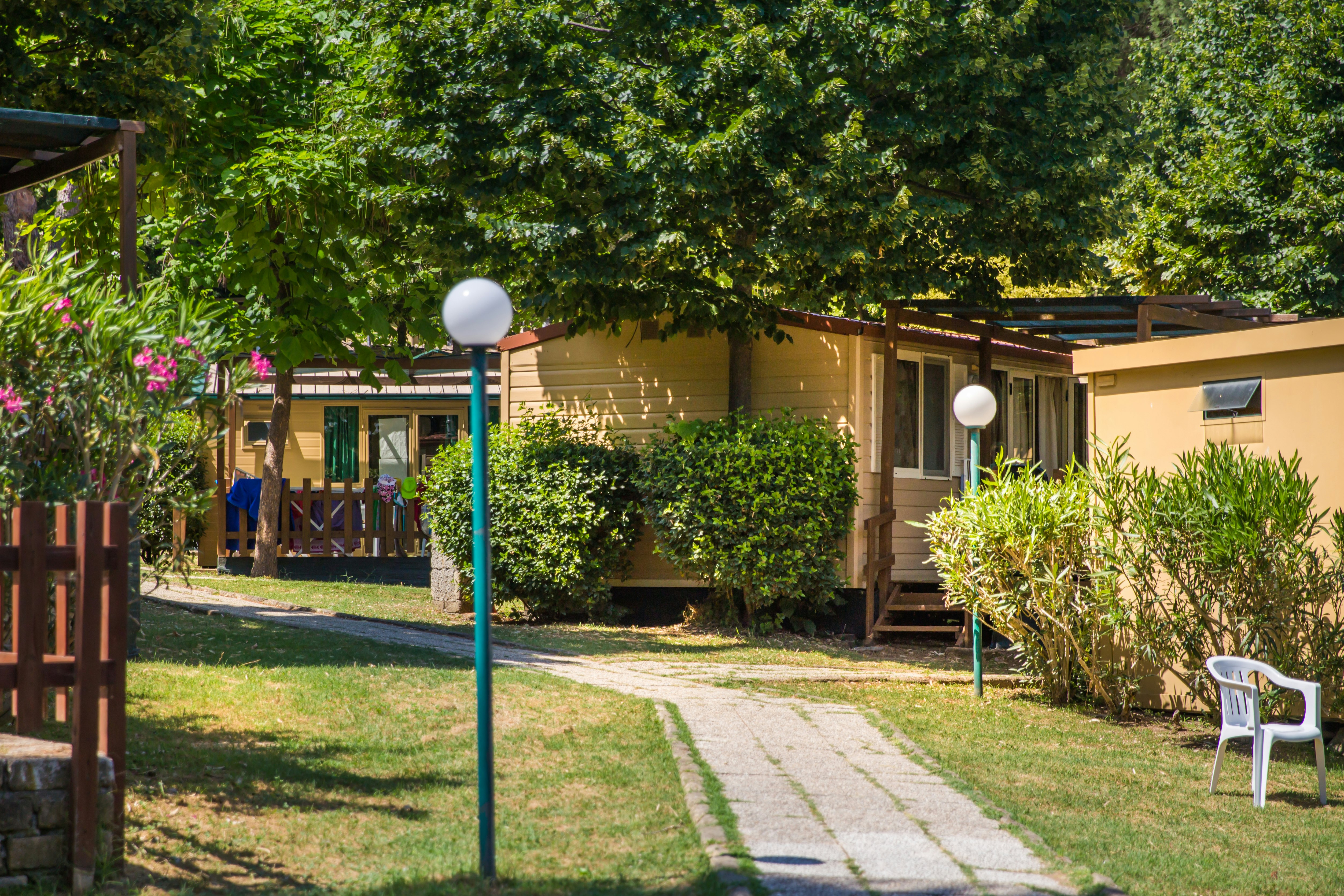 Bungalow im Grünen mit Terrasse, Hecken und Gartenweg, umgeben von Bäumen und Sträuchern.
