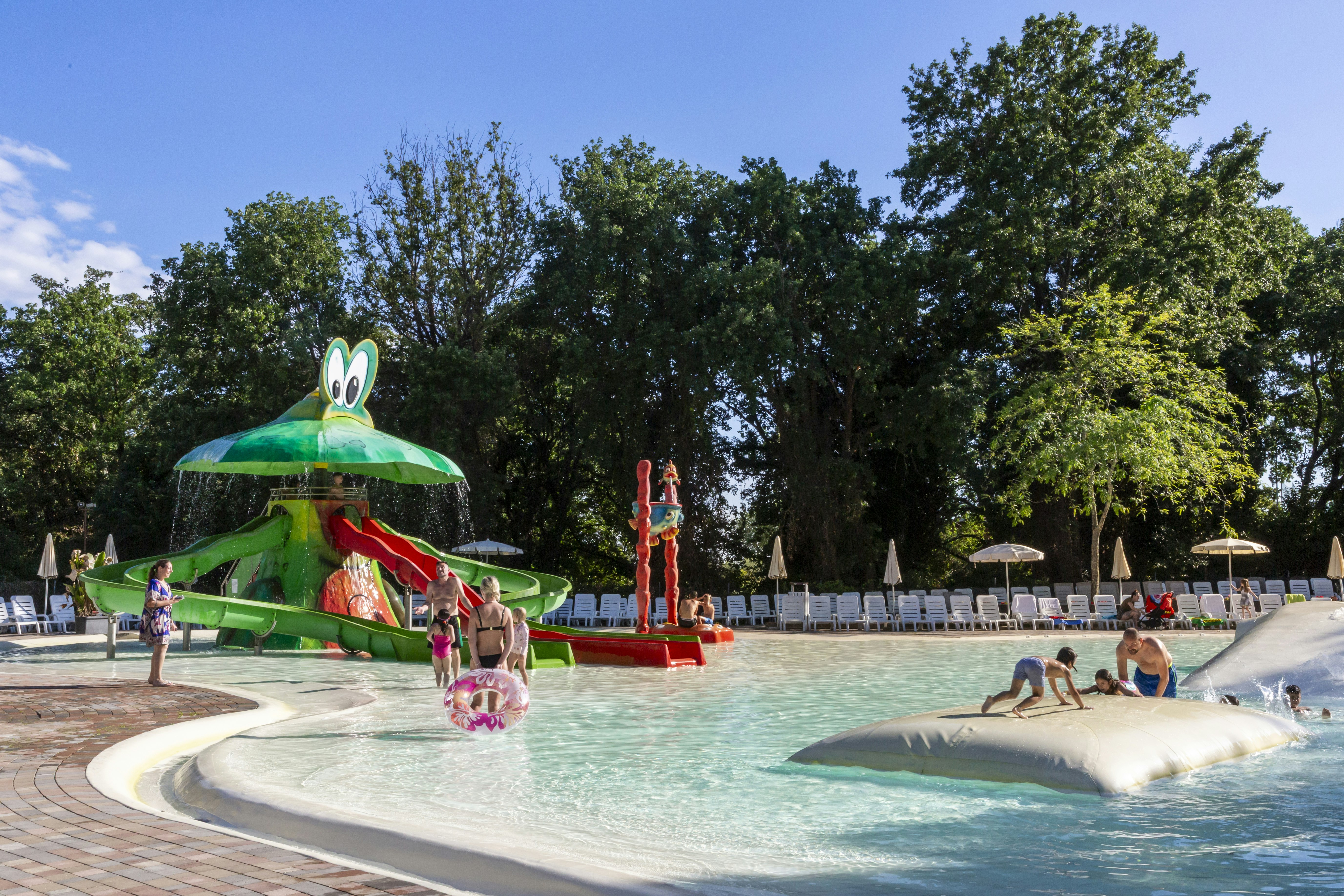 Grüner Wasserrutschen-Spielbereich am Campingpool mit Sonnenschirmen, Liegen und Bäumen im Hintergrund.