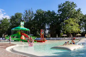Grüner Wasserrutschen-Spielbereich am Campingpool mit Sonnenschirmen, Liegen und Bäumen im Hintergrund.