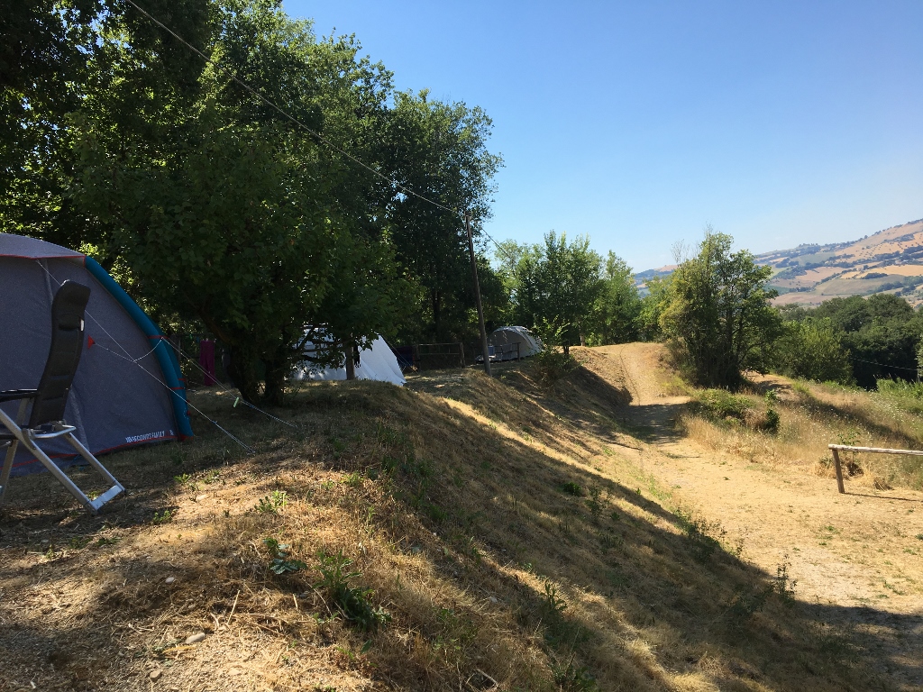 Zelt auf einem grasigen Hang am Campingplatz, daneben ein Weg und Bäume, im Hintergrund Hügel und ein Seeufer.