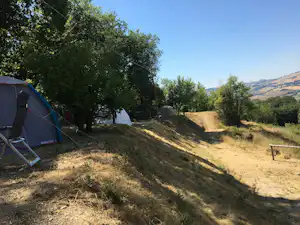 Zelt auf einem grasigen Hang am Campingplatz, daneben ein Weg und Bäume, im Hintergrund Hügel und ein Seeufer.