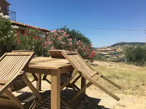 Holzterrasse mit Tisch und Stühlen, blühenden Sträuchern und Blick auf die Hügel im Hintergrund.
