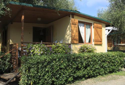 Bungalow mit Terrasse, Holzfensterläden und grüner Hecke, davor ein gepflasterter Weg und Sträucher im Garten.