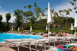 Poolbereich mit Sonnenliegen und weißen Schirmen, im Hintergrund Bäume und ein Blick auf das Gelände des Campingplatzes.