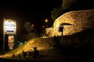 Beleuchteter Campingplatz am Abend: Schild und Weg neben einem steinernen Gebäude mit Rundbogen und Palmen.