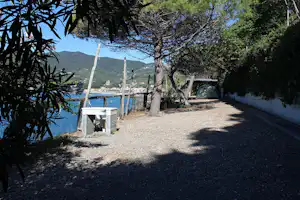 Campingweg am See unter Bäumen, mit Kiesfläche, weißem Zaun und Blick auf das Wasser und die Berge im Hintergrund.