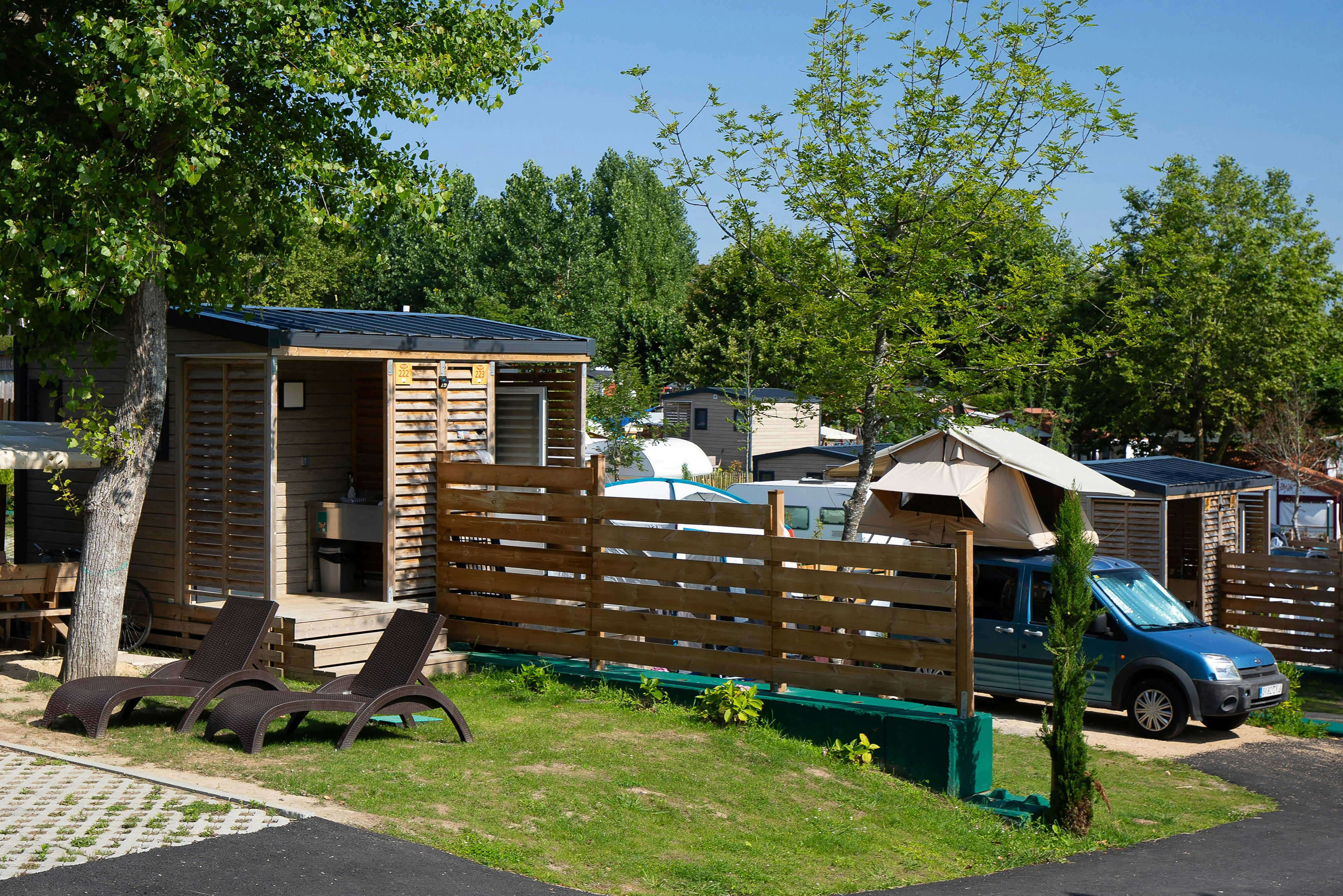 Holz-Mobilheim mit Terrasse, Holzzaun und Liegestühlen auf dem Campingplatz, im Hintergrund Zelte und weitere Fahrzeuge.