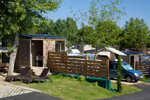 Holz-Mobilheim mit Terrasse, Holzzaun und Liegestühlen auf dem Campingplatz, im Hintergrund Zelte und weitere Fahrzeuge.