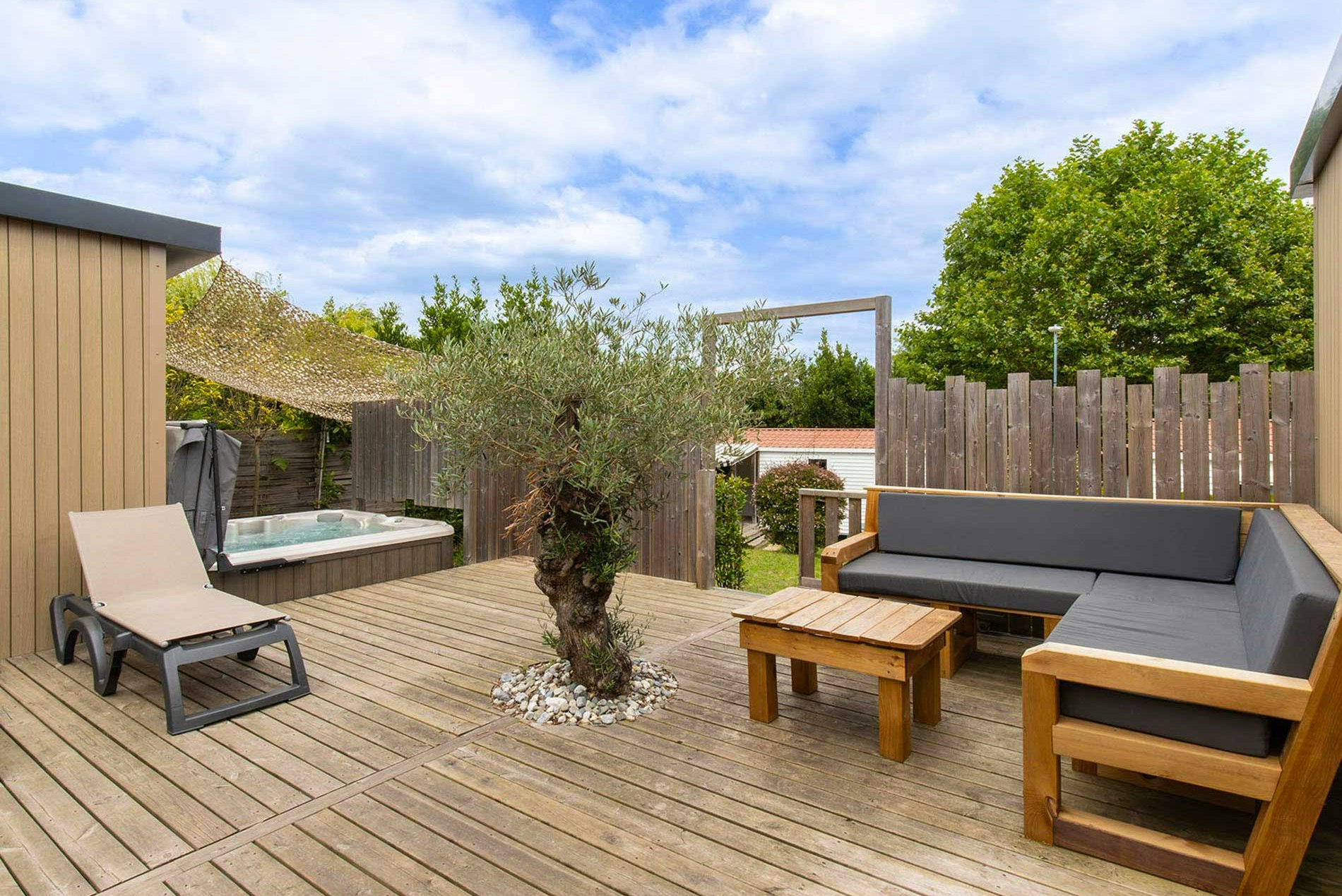 Holzterrasse mit Sitzgruppe, zwei Liegen und kleinem Holztisch, umgeben von Hecken und Sichtschutzzaun.