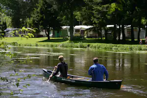 Ruderboot auf dem See vor einem Campingplatz mit Wohnwagen, Zelten und Bäumen am Ufer.