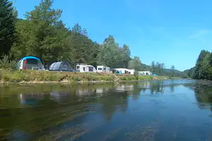 Campingplatz am See: Zelte und Wohnmobile auf einer Wiese, im Hintergrund Bäume und ein ruhiger Uferbereich.