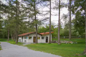 Campinggebäude mit Buro/Ulrich Rezeption, Sitzbank und Stühlen vor dem Eingang, umgeben von Bäumen und Rasen.