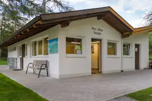 Rezeption am Campingplatz: weißes Gebäude mit Holzvordach, Beschilderung „Büro/Ufficio“ und „Rezeption“, Terrasse mit Bank und Pflasterweg.