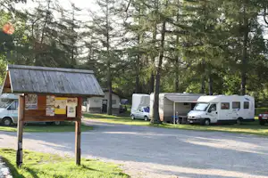 Campingplatz mit Wohnwagen und Campern auf dem Stellplatz, neben einem Holz-Infoboard und Bäumen am Weg.