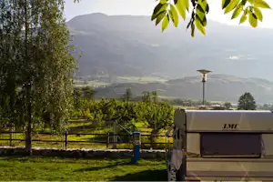Wohnwagen auf einem Campingplatz im Grünen, mit Zaun, Bäumen und Blick auf ein weites Tal in den Bergen.