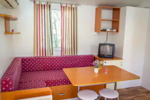 Wohnbereich in einem Campingmobil mit gepolstertem Sofa, Holztisch, zwei Hockern und TV an der Küchenzeile.
