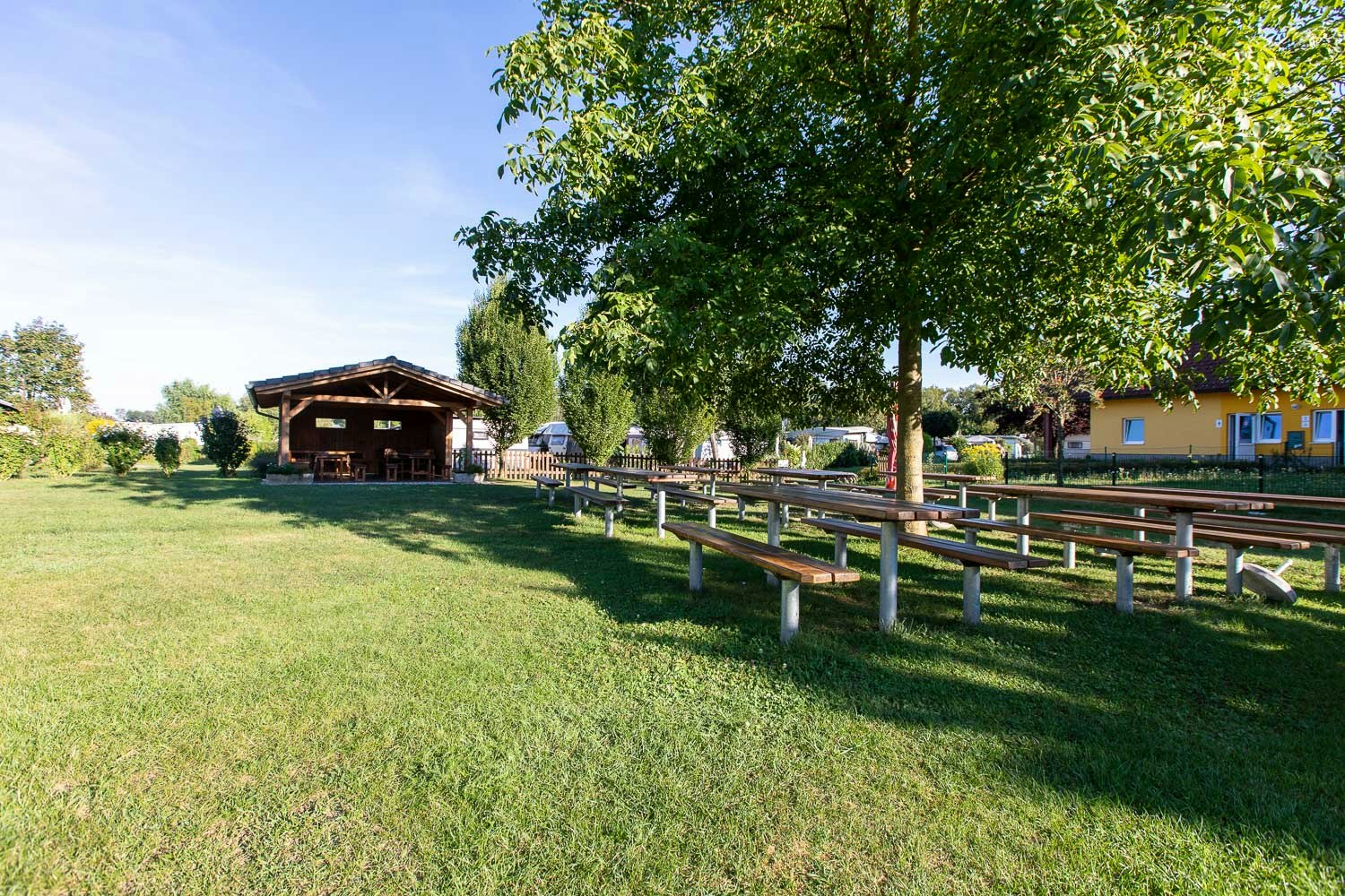 Campingwiese mit Holzpavillon, langen Sitzbänken und Zaun, dahinter Bungalows und Bäume im Grünen.