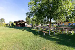 Campingwiese mit Holzpavillon, langen Sitzbänken und Zaun, dahinter Bungalows und Bäume im Grünen.