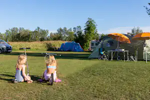 Campingplatz mit zwei Kindern auf dem Rasen, daneben Zelte, Campingstühle und ein Wohnwagen am Stellplatz.