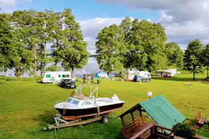 Campingwiese mit Wohnwagen und Boot auf einem Anhänger, Sitzbank und Tisch vor Zelten, umgeben von Bäumen.