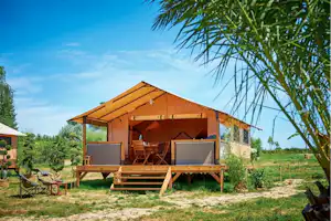 Rustikales Campingzelt auf Holzpodest mit Veranda, Sitzgarnitur und Sonnenschirmen im Garten, umgeben von Bäumen.