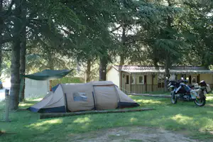 Campingzelt auf dem Stellplatz unter hohen Bäumen, daneben ein Motorrad und ein Holzhaus mit Markise.