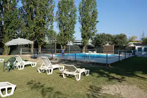 Poolbereich mit blauem Becken, umzäuntem Geländer und Liegestühlen auf dem Rasen, im Hintergrund Bäume und Gebäude.