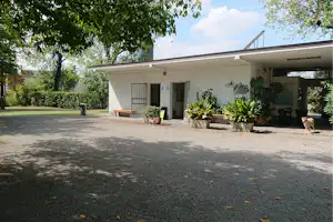 Toiletten-Gebäude auf dem Campingplatz mit Eingangstüren, Pflanzenkübeln und Sitzbank vor gepflastertem Weg.