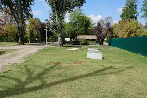 Grüne Campinganlage mit Rasen, Kiesweg, Bäumen und einer Bank am Rand einer grünen Sichtschutzwand.
