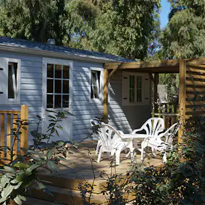 Bungalow mit Terrasse und weißen Gartenstühlen, Holzüberdachung und Pflanzen im Vordergrund, umgeben von Bäumen.