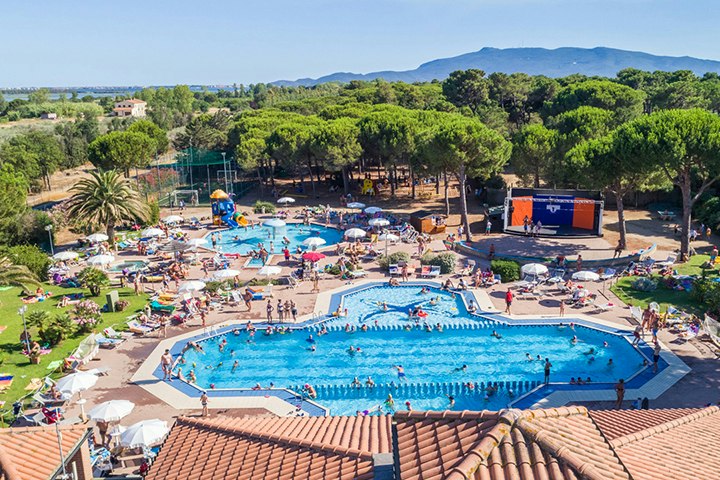 Großes Camping-Schwimmbecken mit vielen Liegestühlen, Sonnenschirmen und Menschen, umgeben von Bäumen und Hügeln im Hintergrund.