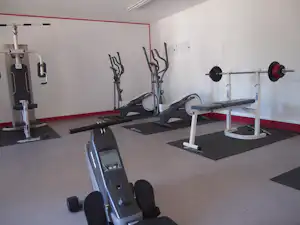 Fitnessraum im Campingbereich mit Trainingsgeräten, Hantelbank und Langhantelstange an der Wand.