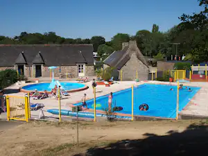 Camping-Poolanlage mit blauem Becken, gelben Absperrgittern und mehreren Ferienhäusern aus Stein im Hintergrund.