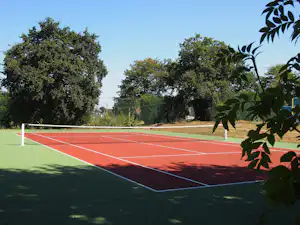Roter Tennisplatz auf grünem Rasen, umgeben von Bäumen und Hecken, mit Zaun im Hintergrund.