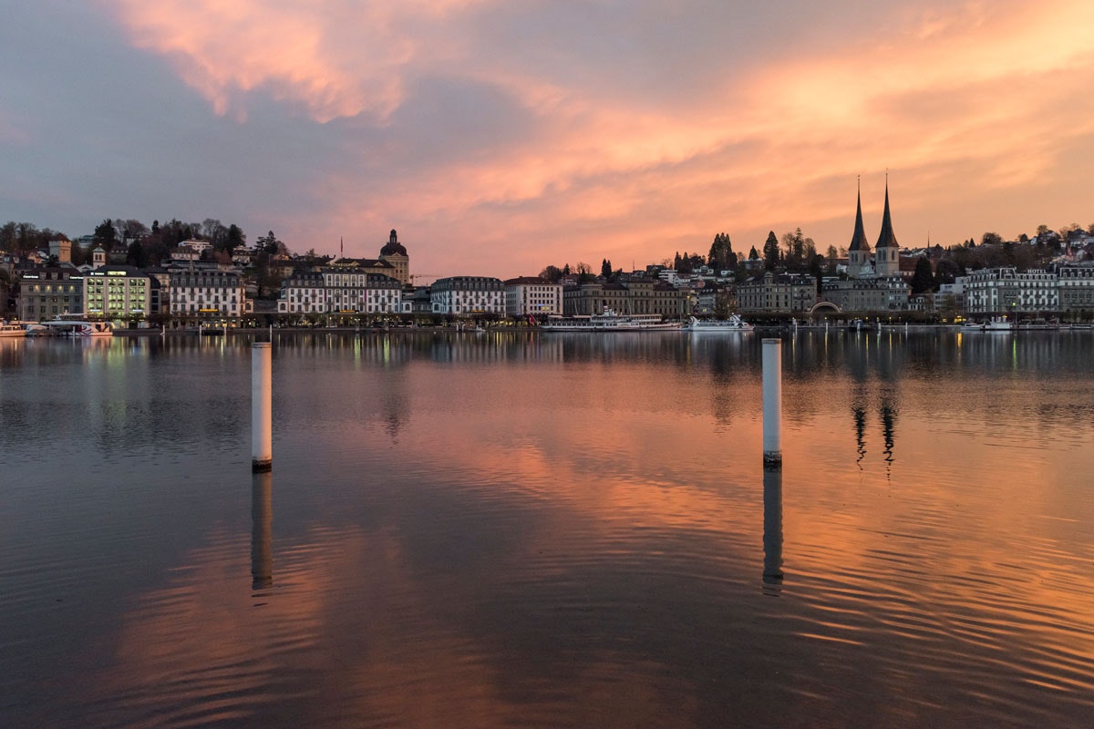 Spiegelnde Uferpromenade am See bei Sonnenuntergang, im Hintergrund Häuser und eine Kirchturmspitze.