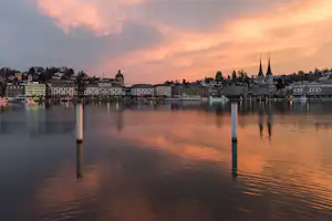 Spiegelnde Uferpromenade am See bei Sonnenuntergang, im Hintergrund Häuser und eine Kirchturmspitze.
