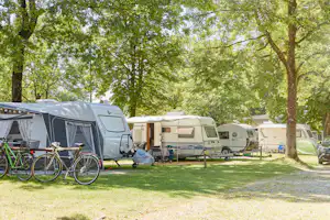 Wohnmobile und Zelte auf einem Campingplatz unter Bäumen, mit Fahrrädern am Stellplatz und Rasenfläche.
