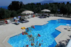 Camping-Pool mit blauem Wasser, Liegen und Sonnenschirmen auf der Terrasse, im Hintergrund Bäume und Meerblick.