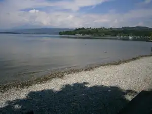 Seeufer mit Kiesstrand, ruhigem Wasser und Blick auf Uferpromenade, Bäume und Hügel in der Ferne.