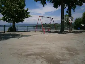 Spielplatz mit roter Schaukel auf Betonfläche am See, Bäume und Geländer am Ufer im Hintergrund.